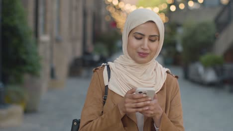 Muslim-Woman-Wearing-Hijab-Walking-Along-City-Street-Sending-Text-Message-To-Date-On-Mobile-Phone-1